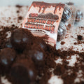 A pack of Protein Buff Pops - Death By Chocolate sits atop crumbled chocolate, with several protein-packed chocolate balls arranged in front, creating a decadent dessert scene that evokes the richness of chocolate cake—ideal for an on-the-go treat.