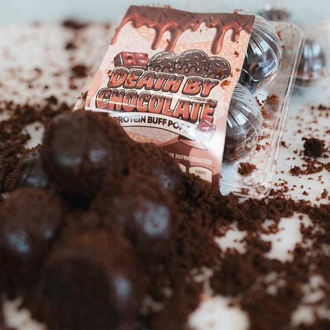 A pack of Protein Buff Pops - Death By Chocolate sits atop crumbled chocolate, with several protein-packed chocolate balls arranged in front, creating a decadent dessert scene that evokes the richness of chocolate cake—ideal for an on-the-go treat.
