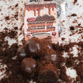 A package of Protein Buff Pops - Death By Chocolate rests among scattered chocolate crumbs, with several round chocolate cake treats in front—ideal for a quick grab-and-go protein boost.