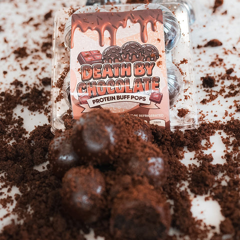 A package of Protein Buff Pops - Death By Chocolate rests among scattered chocolate crumbs, with several round chocolate cake treats in front—ideal for a quick grab-and-go protein boost.