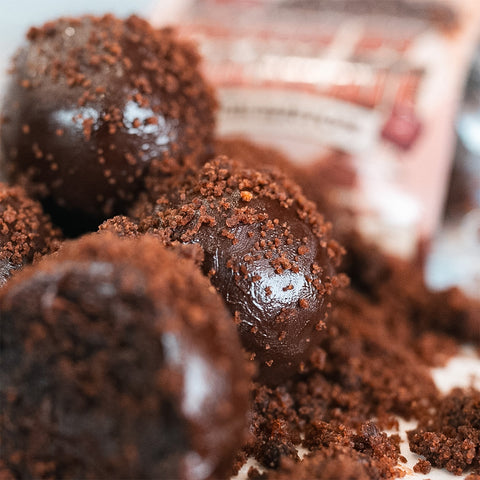 Close-up of Protein Buff Pops - Death By Chocolate, decadent truffle bites coated in chocolate crumbs with more crumbs scattered around—each bite bursting with the rich flavor of chocolate cake. A blurred package of Protein Buff Pops - Death By Chocolate appears in the background, making these treats perfect for a quick grab-and-go indulgence.