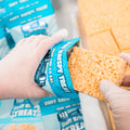Wearing gloves, someone puts a Buff Krispy Treat protein snack into a blue Buff Krispy Treat bag, with more treats and packaging in the background—ideal for muscle recovery.