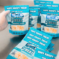 A gloved hand holds a Buff Krispy Treat Original Krispy above identical Buff Krispy Treat snack bags on a counter, each featuring "Grab and Go Bag" at the top—perfect for quick muscle recovery.