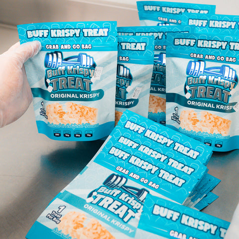 A gloved hand holds a Buff Krispy Treat Original Krispy above identical Buff Krispy Treat snack bags on a counter, each featuring "Grab and Go Bag" at the top—perfect for quick muscle recovery.