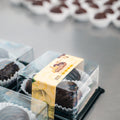 Close-up of 3 Banana Chocolate Protein Poppers, a healthy snack in clear plastic boxes with yellow labels on a metal surface; more high protein chocolates in white paper cups are blurred in the background.