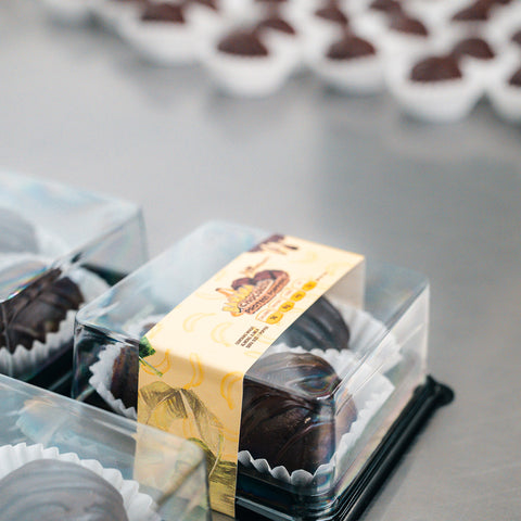 Close-up of 3 Banana Chocolate Protein Poppers, a healthy snack in clear plastic boxes with yellow labels on a metal surface; more high protein chocolates in white paper cups are blurred in the background.