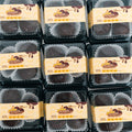 Plastic containers of 3 Banana Chocolate Protein Poppers are arranged in a grid. Each has a beige label with chocolate graphics, and the high-protein treats inside are served in white paper cups for a healthy snack option.