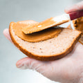 A gloved hand holds a slice of bread as a spatula spreads Peanut Butter Jelly Sandwich filling, making a high-protein snack perfect for post-workout recovery.