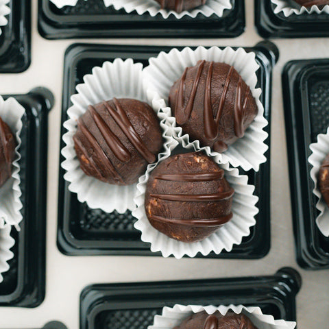Close-up of 3 Banana Chocolate Protein Poppers in white paper liners on a black tray. These round treats are chocolate-drizzled, high-protein, and more trays of this healthy snack appear in the background.