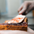 A close-up shows a gloved hand spreading strawberry jam onto bread with a spatula, preparing a Peanut Butter Jelly Sandwich packed with protein—ideal for your workout sandwich routine.