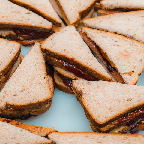 A close-up of several Peanut Butter Jelly Sandwiches, protein-packed and cut into triangles, arranged in a circular pattern on whole wheat bread over a white surface.
