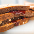 A close-up of the Peanut Butter Jelly Sandwich, made with whole grain bread and packed with protein, shows creamy peanut butter and red jelly layered between each slice.