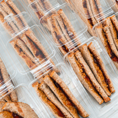 Rows of clear plastic containers, each with two halves of a Peanut Butter Jelly Sandwich made with whole wheat bread and packed with protein, are neatly arranged on a light surface.
