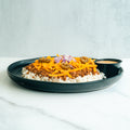 A black plate with white rice topped with seasoned ground meat, shredded cheddar cheese, diced red onions, and jalapeño slices. A small black cup of sauce is on the side. The background is a light marble surface.