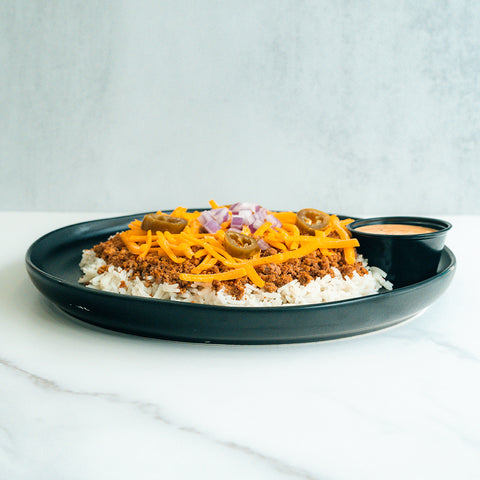 A black plate with white rice topped with seasoned ground meat, shredded cheddar cheese, diced red onions, and jalapeño slices. A small black cup of sauce is on the side. The background is a light marble surface.