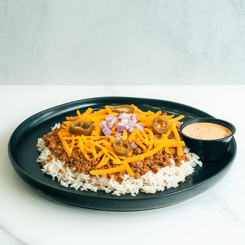 A black plate with white rice topped with seasoned ground meat, shredded cheddar cheese, diced red onions, and sliced jalapeños. A small black cup of creamy dipping sauce is on the side.