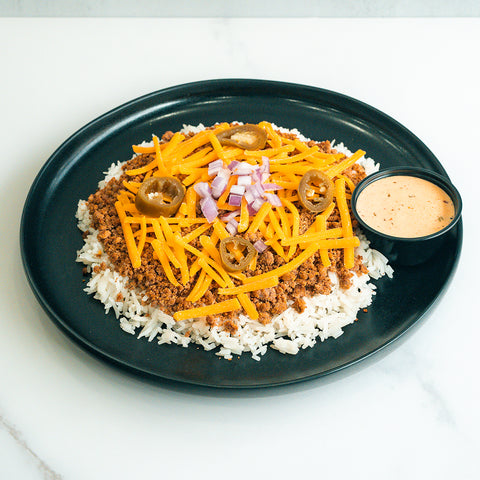 A black plate with white rice topped with ground meat, shredded cheddar cheese, sliced jalapeños, and chopped red onions, served with a small cup of creamy dipping sauce on the side.