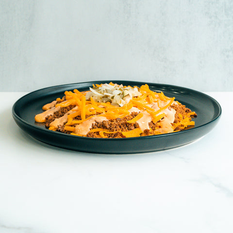 A black plate holds a serving of food with ground beef, shredded cheddar cheese, chopped onions, and a drizzle of creamy orange sauce on top, set against a white background.