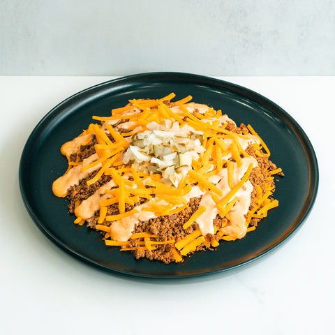 A black plate with seasoned ground beef, shredded cheddar cheese, diced onions, chopped pickles, and a drizzle of creamy orange sauce, arranged in layers.