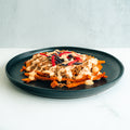 A black plate holds a serving of loaded sweet potato fries topped with ground meat, creamy sauce, and colorful tortilla strips, set against a light background.