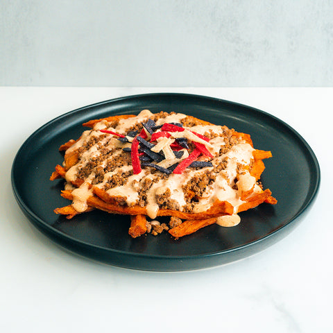 A black plate with sweet potato fries topped with ground beef, creamy sauce, and colorful tortilla strips, set against a light background.
