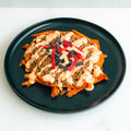A black plate with sweet potato fries topped with ground meat, creamy sauce, and colorful tortilla strips, on a white surface.