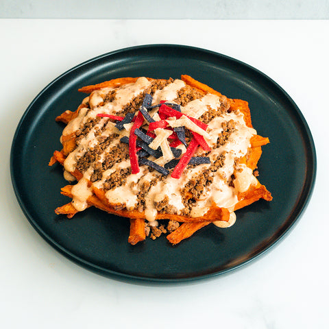 A black plate with sweet potato fries topped with ground meat, creamy sauce, and colorful tortilla strips, on a white surface.
