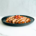 A black plate with cubed, seasoned potatoes topped with a creamy sauce, crumbled bits, and a fresh tomato salsa, set on a white surface with a light background.