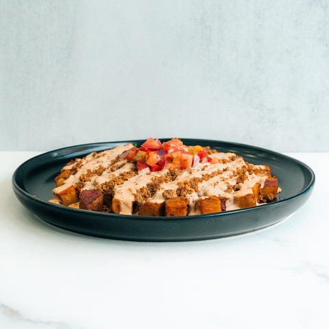 A black plate with cubed, seasoned potatoes topped with a creamy sauce, crumbled bits, and a fresh tomato salsa, set on a white surface with a light background.