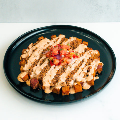 A black plate with crispy cubed potatoes topped with crumbled ground meat, drizzled with a creamy sauce, and garnished with a scoop of fresh pico de gallo in the center.