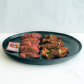 The Garlic Flank Bowl features spiced grilled chicken, purple roasted potatoes, and a packet of spicy ketchup, all served on a black plate to create a vibrant, high-protein meal set against a white background.