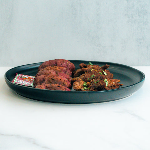 The Garlic Flank Bowl features sliced cooked beef and shredded meat garnished with herbs, served with hot sauce on a black plate atop a white marble surface against a light background.