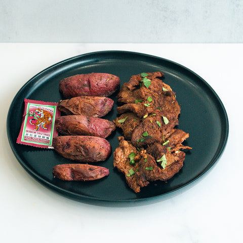 The Garlic Flank Bowl features sliced roasted purple sweet potatoes, garlic flank steak pieces topped with herbs, and a side of sriracha hot sauce—a flavorful high-protein meal.