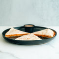 Three Buffalo Chicken Quesadilla slices sit on a round black plate with a small cup of dipping sauce—set on a white surface and light gray background, this makes for an ideal high-protein meal prep option.