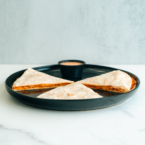 Three Buffalo Chicken Quesadilla slices sit on a round black plate with a small cup of dipping sauce—set on a white surface and light gray background, this makes for an ideal high-protein meal prep option.