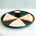 Three Buffalo Chicken Quesadilla wedges are served on a round black plate with dipping sauce in the center, set on a white surface with a light gray background—a perfect high-protein meal for easy prep.