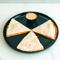 The Buffalo Chicken Quesadilla features three wedges arranged in a circle on a black plate with a small cup of orange dipping sauce, ideal for meal prep and served on a white surface.