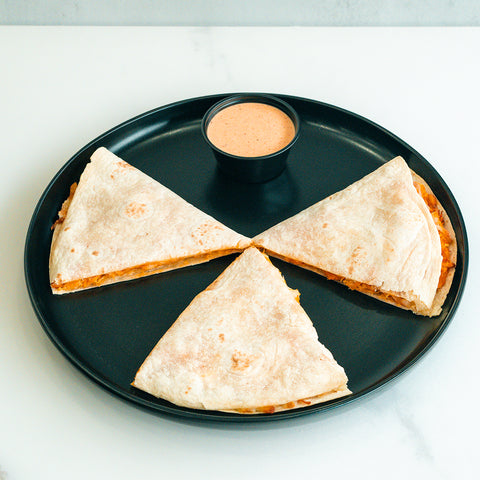 The Buffalo Chicken Quesadilla features three wedges arranged in a circle on a black plate with a small cup of orange dipping sauce, ideal for meal prep and served on a white surface.