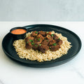 Enjoy “GLORIOUS GAINS” Steak Bites Cilantro Lime Rice—a black plate of tender steak bites over seasoned cilantro lime rice, garnished with herbs, plus a zesty orange dipping sauce. Perfect for high protein meal prep.