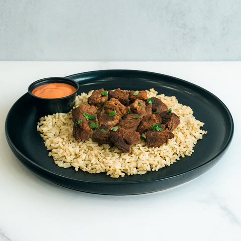 Enjoy “GLORIOUS GAINS” Steak Bites Cilantro Lime Rice—a black plate of tender steak bites over seasoned cilantro lime rice, garnished with herbs, plus a zesty orange dipping sauce. Perfect for high protein meal prep.