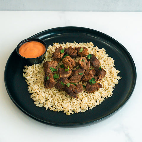 Enjoy “GLORIOUS GAINS” Steak Bites Cilantro Lime Rice—a high-protein meal with tender steak bites on cilantro lime rice, garnished with herbs, and served alongside a zesty orange dipping sauce. Perfect for easy meal prep.