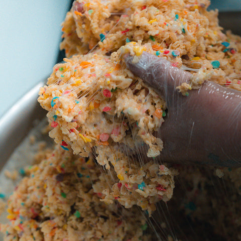 A gloved hand mixes a sticky, colorful cereal and marshmallow mixture in a large metal bowl to make a Fruity Cereal Buff Cereal Bar.