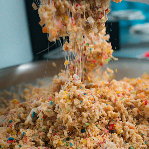 A close-up of Fruity Cereal Buff Cereal Bar treats being mixed with melted marshmallows, gooey strands stretching as the protein-packed fruity cereal mixture is lifted from a large metal bowl.