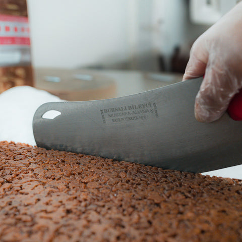 A gloved hand uses a large metal knife with engraved text to slice through a dense tray of Chocolate Buff Cereal Bar, loaded with chopped nuts—a hearty snack perfect for the gym.