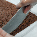 A gloved hand slices a Chocolate Buff Cereal Bar—a rectangular chocolate protein snack similar to a rice crispy treat—on a white surface, making it an ideal gym snack.