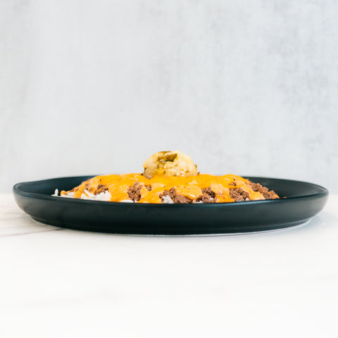 A black plate showcases the Cheeseburger Bowl—gluten-free rice topped with ground beef, melted cheddar cheese, and guacamole—presented against a light, neutral background.