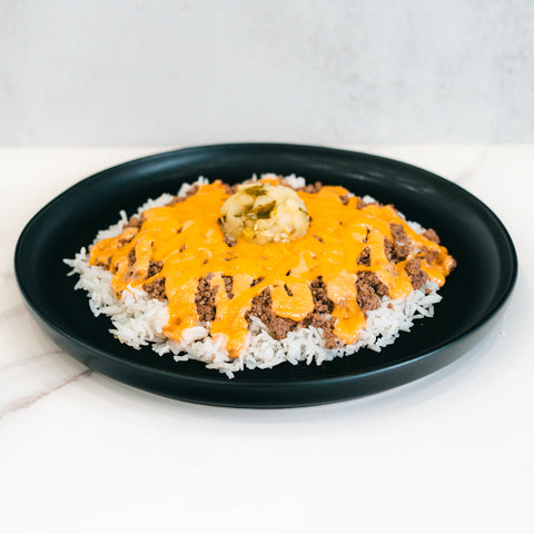 The Cheeseburger Bowl is served on a black plate with white rice, savory ground beef, orange cheese sauce, mashed potatoes, and green relish at the center. It’s gluten-free.