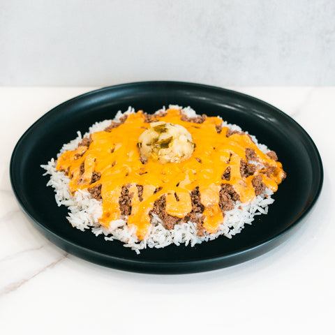A black plate features the Cheeseburger Bowl: white rice with ground beef, melted orange cheese sauce, mashed potatoes in the center, and fresh green jalapeño slices on top. Gluten-free.