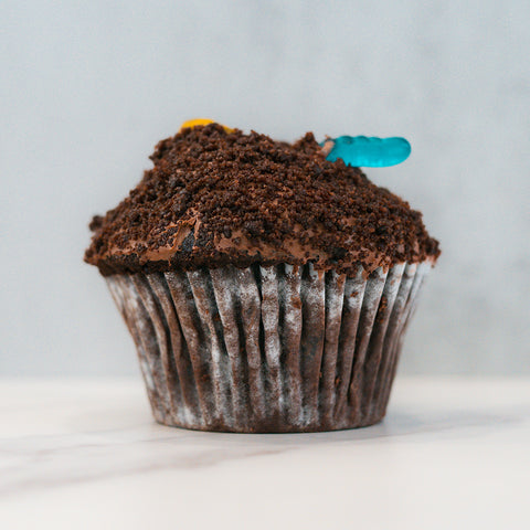 Buffin Muffin Dirt Cake is a chocolate muffin topped with crumbled chocolate cookie crumbs and a playful gummy worm, designed to look like dirt with a worm. Bite inside to reveal a rich chocolate filling for an extra sweet surprise.