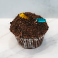 The Buffin Muffin Dirt Cake features a chocolate cupcake filled with rich chocolate, topped with crushed cookie crumbs and two gummy worms—one yellow and one blue—arranged on a white marble surface.